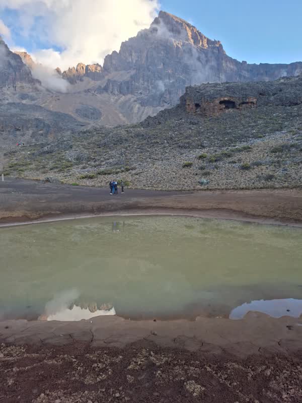 Mawenzi Tarn at 4,330 m, the jagged peak mirrored in the shallow green lake with two trekkers on the shore path.