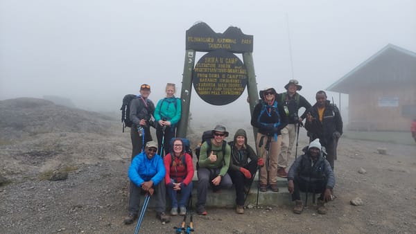 Trekkers and crew at the carved Shira II Camp signboard, 3,850 m, in moorland mist on the Shira Plateau crossing shared by the Machame and Lemosho routes.