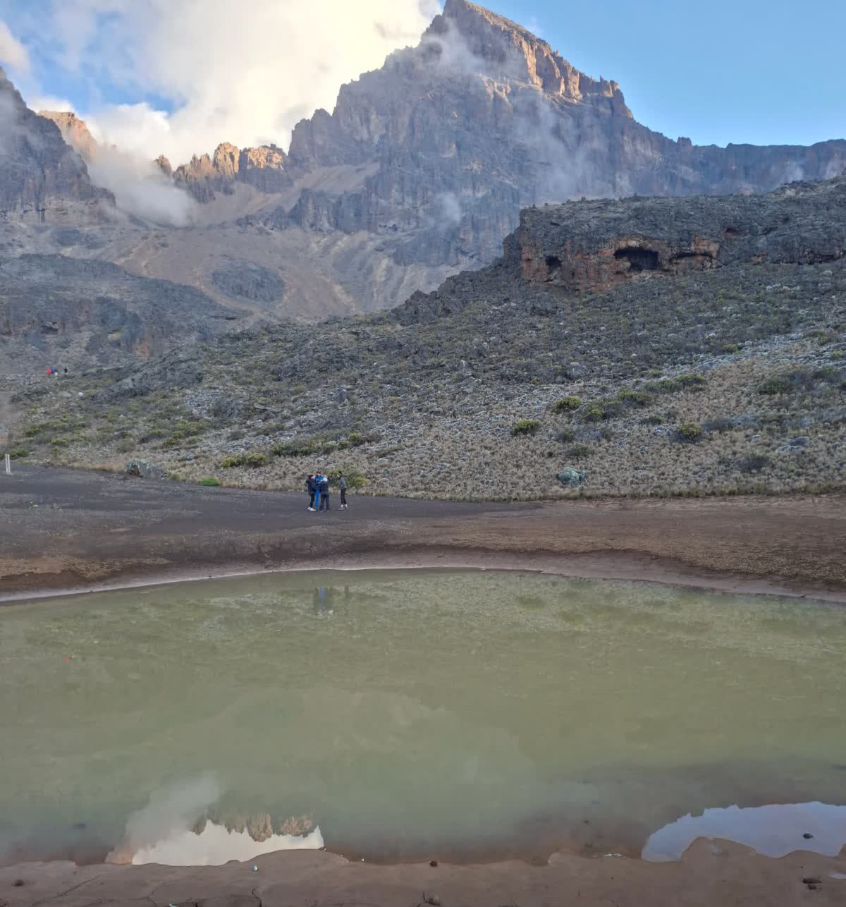 Mawenzi Tarn at 4,330 m, a shallow green lake reflecting the jagged Mawenzi peak, with two trekkers on the shoreline path