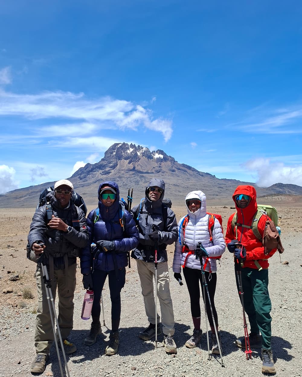 Five guides and climbers with trekking poles and packs standing together on the gravel plain of the Saddle, the jagged Mawenzi Peak rising behind them under a blue sky