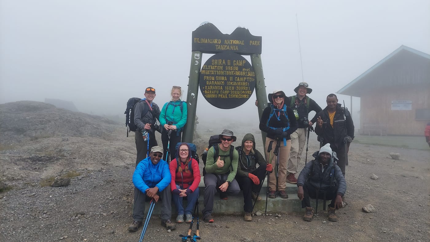 Trekkers and crew grouped around the carved Shira II Camp signboard at 3,850 m in moorland mist
