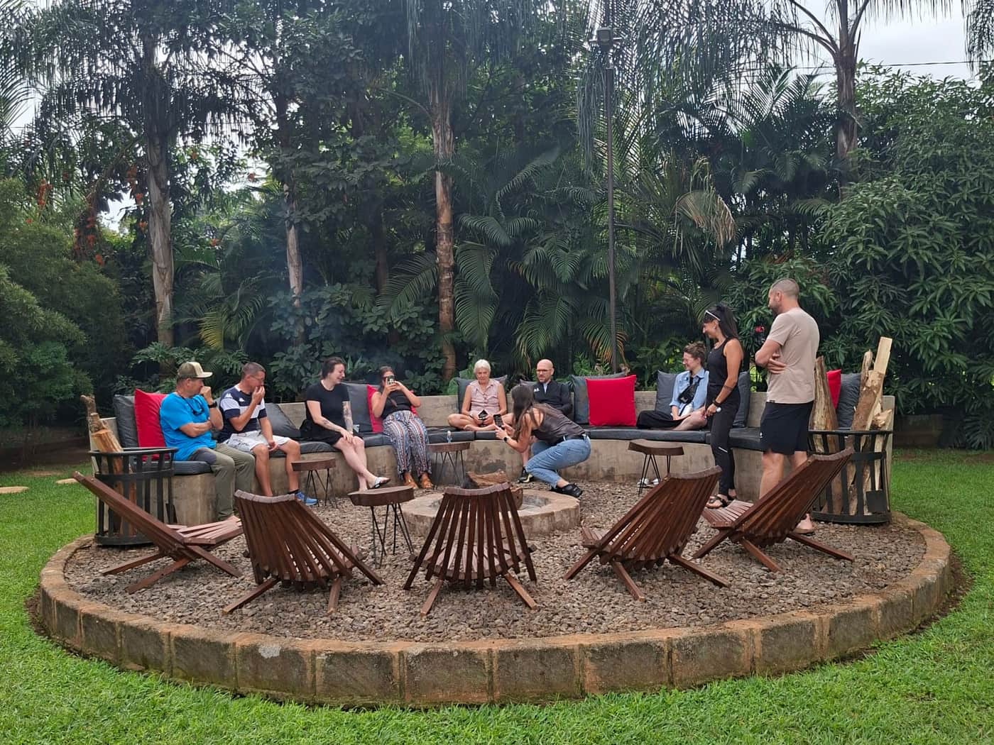 Climbers sitting around the stone fire-pit circle in the palm garden of the Moshi base, wooden chairs set out on the gravel and lawn around them