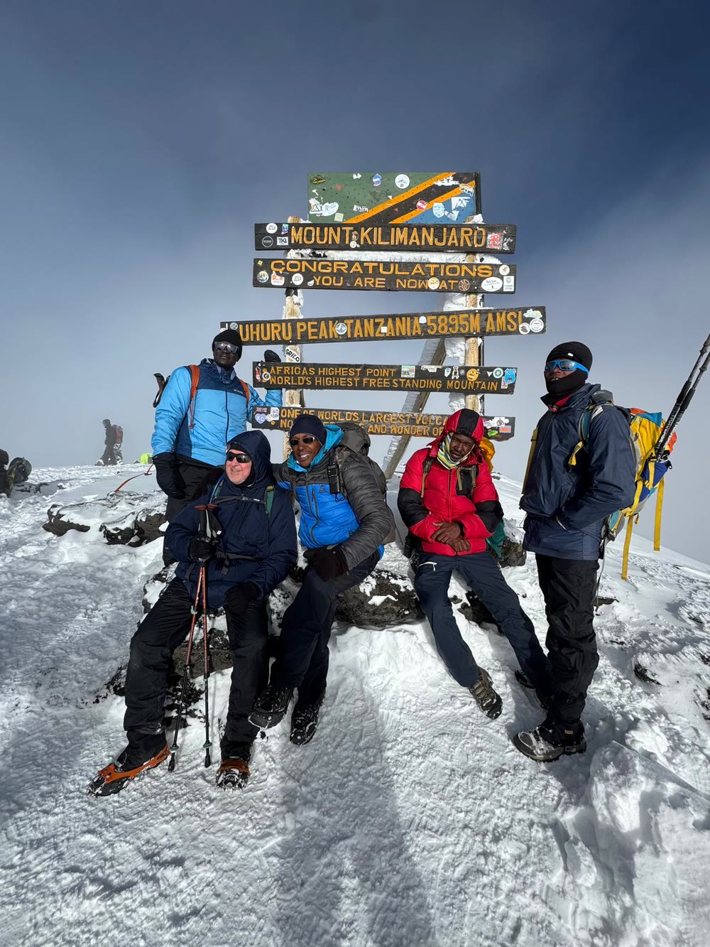 Nelson Mushi, seated in the blue jacket, with his climbers and guides at the carved Uhuru Peak signboard reading Tanzania 5,895 m AMSL, Africa's highest point, world's highest free standing mountain, in snow and blowing cloud on the summit