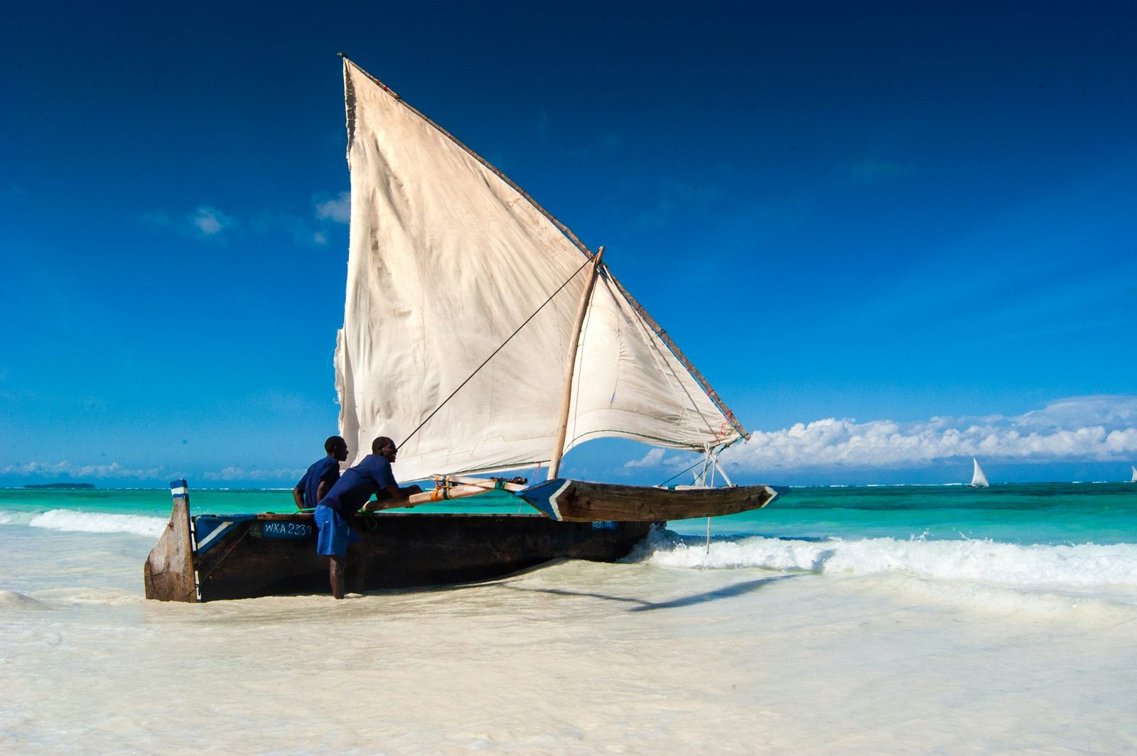 A traditional wooden Zanzibari dhow with its sail raised on white sand at the turquoise water's edge, two local sailors beside it and another dhow on the horizon